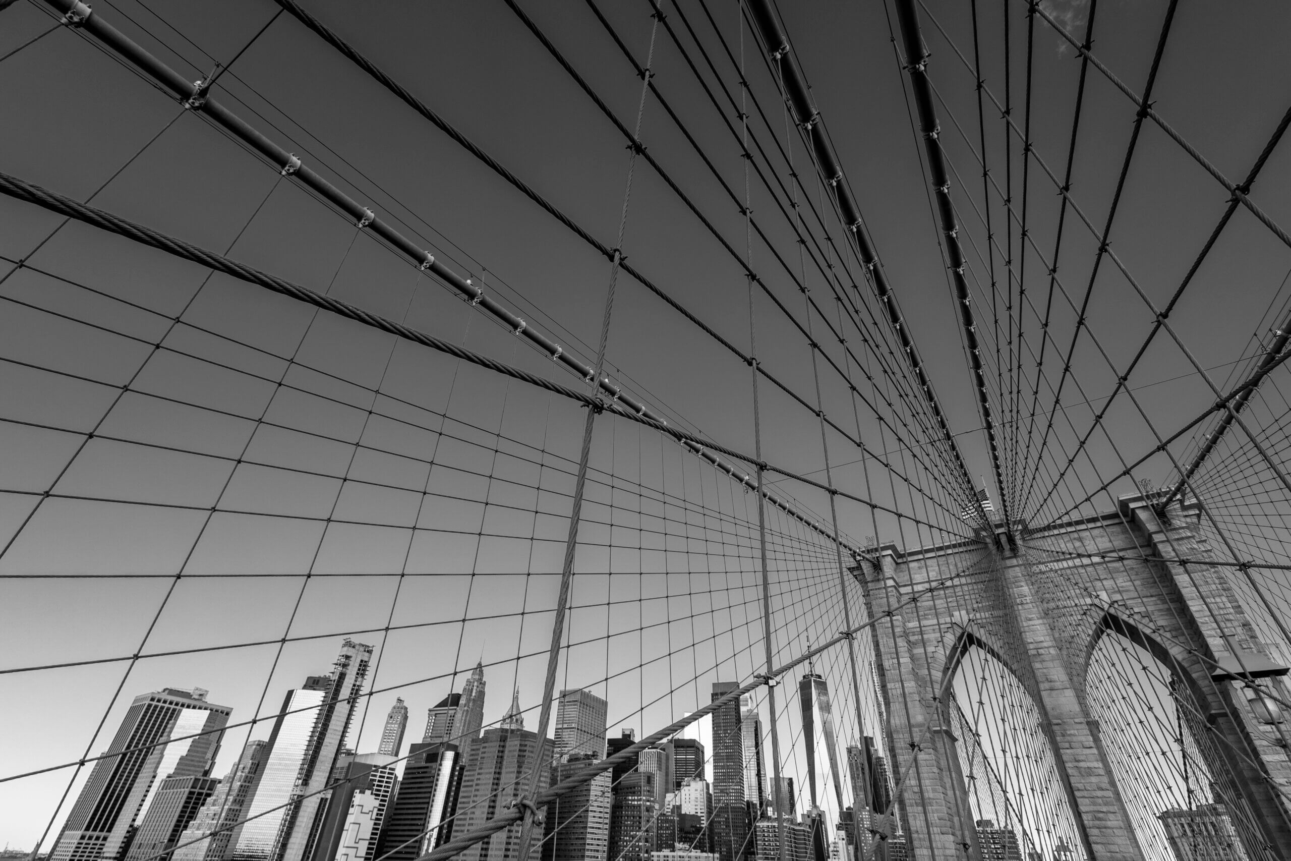 A black-and-white photo capturing the iconic cables of the Brooklyn Bridge, with the Manhattan skyline and its skyscrapers visible in the background