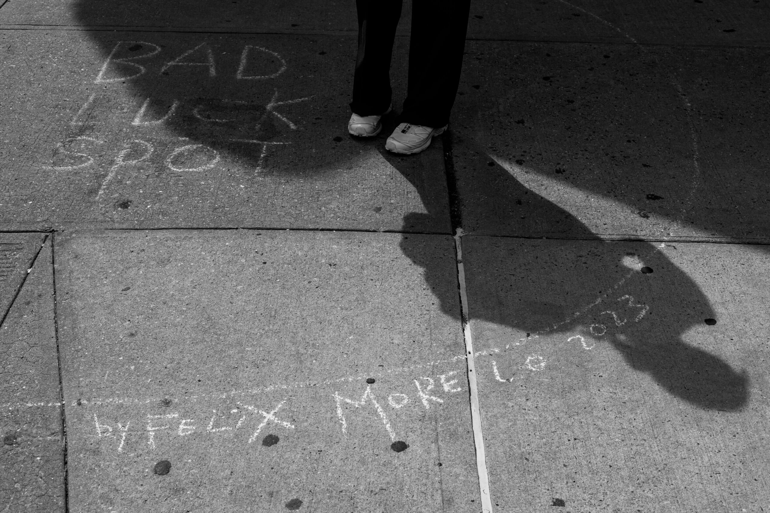 A black-and-white photo of Félix Morelo's street art in New York, showing a circle on the ground labeled "Bad Luck Spot" with a woman's feet inside the circle