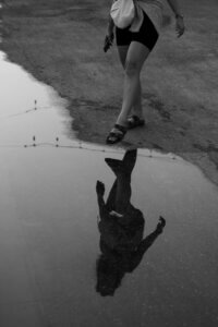 A black-and-white photo of a rain puddle in Hoi An, Vietnam, reflecting the silhouette of a woman miming walking in the water