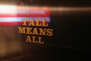 A restroom door at the Rodeo Bar in Dallas with the phrase “Y’all Means All” overlaid on a transparent American flag, symbolizing gender inclusivity