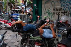 Two men reclining on their motorcycles in Vietnam, napping while playing on their smartphones